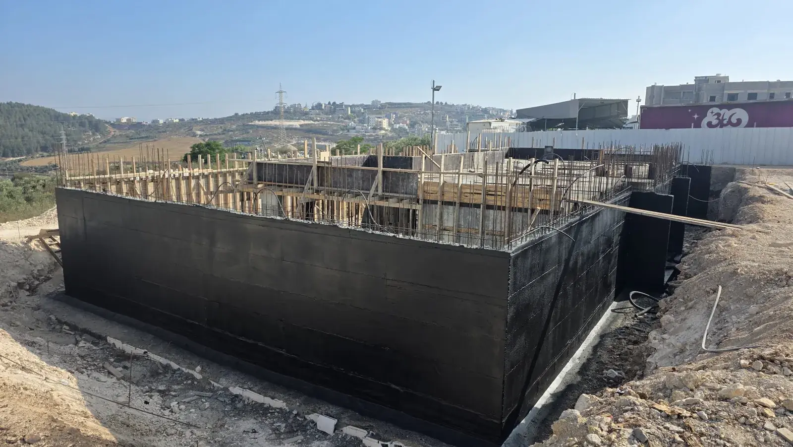 קירות יסוד של מבנה בבנייה מצופים בשכבת איטום שחורה, עם ברזלי זיון וטפסות עץ מעליהם